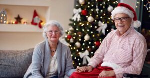 Elderly couple celebrating Holidays At Home by a Christmas tree, holding hands and smiling.
