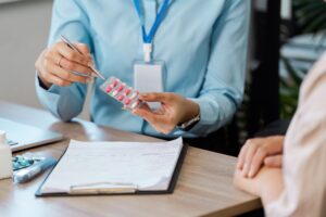 Nurse reviewing medication management with Hospice Patient, the nurse is sitting down with patient at table while holding the pills and a pen. Nurse also has a clipboard and laptop on the table.