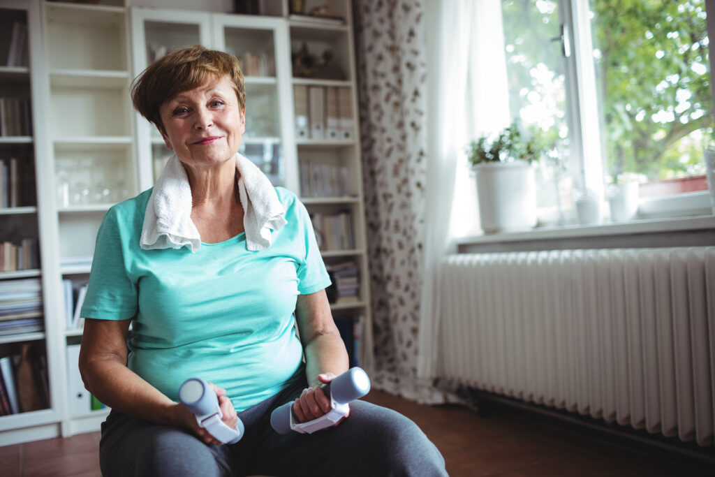 Senior woman exercising with dumbbells at home, towel around neck. Exercising for seniors.