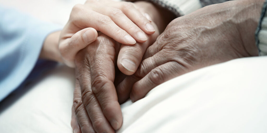 Close-up of a younger hand holding an older, wrinkled hand on a white bed. Shows care and support.