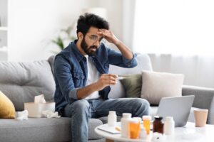 Man with fever checks thermometer while working from home. Medication management is important when sick.