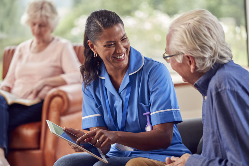 Caregiver showing tablet to senior man, with another senior woman in the background. Hospice care setting.